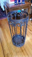Photo of ornate metal umbrella stand with scrollwork design, cylindrical shape, and dark metal finish on wood floor.