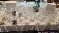 Full view of wine glasses arranged on a lace tablecloth with two white boxes and a small silver cup, totaling approximately 18 glasses of various shapes and sizes.