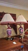 Pair of matching ceramic base table lamps with floral designs, fabric pleated lampshades, placed on a wooden surface against a beige wall.