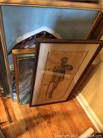 Stack of six framed and canvas art pieces leaning against a wall showing different frame styles and damages