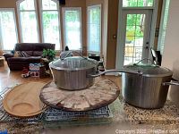 Photo of Calphalon covered pots, Pampered Chef pizza stone, cutting boards and wire cooling racks on a wooden table with living room background.