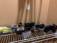 All items displayed on shelf including Canon AV-1 camera set, teleconverter, flash, Pentax film camera, charger.