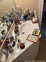 Overview of multiple cat figurines displayed on a white countertop including different sizes and colors with two framed cat pictures on the side.