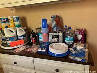 Wide view of countertop with cleaning supplies, paper plates, cups, light bulbs, and batteries, showing overall contents of lot.