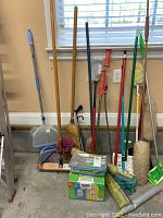 Wide shot of multiple cleaning supplies including brooms, mops, dusters, plumbing snake, cleaning wipes and dustpans neatly arranged against a wall.