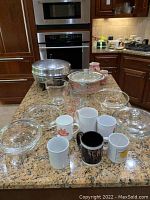 Full lot of glass cookware, measuring cups, pitchers, coffee mugs, and Farberware electric fry pan on kitchen counter.