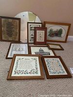 Overview photograph showing nine framed embroidery artworks arranged leaning against a wall and floor, along with one large round mirror with mosaic frame.
