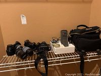 Overview of all items on white wire shelf: cameras, lenses, charger in foam block, handheld radio, camera bag
