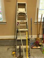 Full view of 6 foot aluminum All American brand step ladder with 5 steps, along with measuring tools, grabber and reflective markers on the floor next to it