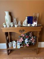 Wooden table with heart-shaped cutout, decorated with various figurines, glass vases, mugs, a silver tray, and faux flower arrangement underneath.