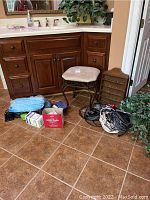 Photo showing a wooden bathroom cabinet with various items on the floor including pill organizer, cables, faux plants, and a padded stool.