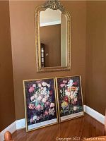 Full view of the Victorian style wall mirror hanging on the wall above two floral framed prints sitting on the floor.