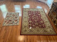 Two rugs displayed on wood floor: larger 5x8 burgundy and tan area rug next to smaller 4x2 floral welcome mat in burgundy and blue.