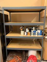 Full view of metal framed utility shelf with particleboard shelves holding paint cans, brushes, and a tray.