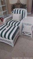 Full view of white wicker chair with vertical green and white striped cushions, a pillow, matching ottoman, and side table on carpeted floor.