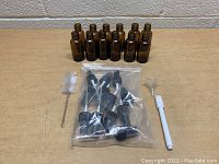 12 small amber glass spray bottles, spray tops in plastic bag, small white pen with brush tip, small brush cleaner, and clear plastic funnel placed on a table.
