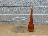 Clear pressed glass compote vase and tall amber art glass bud vase photographed side by side on a wooden surface with a light gray brick background.