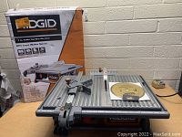 Ridgid 7-inch tile saw on a wood table next to the original box, showing the front and top of tool, diamond blade installed under blade guard.