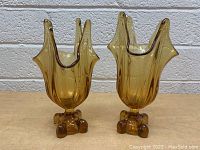 Front view of two amber glass vases with tall, wavy, flame-like rims and rounded textured bases, placed side by side on a tan surface against a white brick wall.