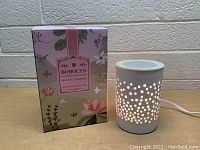 White ceramic Bobolyn wax melt warmer shown next to its floral-themed retail box. The warmer features a pattern of small round holes for light diffusion; electric cord attached.