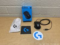 Lot contents displayed on wooden surface against beige brick wall: Logitech G300S wired gaming mouse, original blue retail box, mouse bungee, and two Logitech logo stickers.