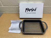 Parini rectangular cast iron grill dish with original box and instruction sheet placed on a wooden surface against a beige brick wall.