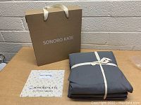 Sonoro Kate gift bag in kraft paper with cream handles, dark grey bed sheets tied with ribbon, and Sonoro Kate thank you card on a table.