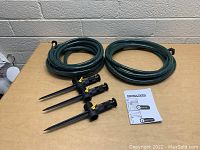 Three black spike sprinklers, two green garden hoses, and instruction manual on a wooden table against a brick wall.