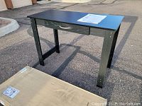 Black writing desk standing assembled outdoors with original packaging box nearby on pavement.