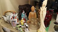 Collection of Asian themed figurines and teapot on shelf, showing wood, ceramic, resin, glass, and stone items.