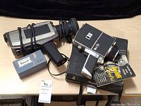 Toshiba video camera with Nikkor zoom lens, DeJUR Super 8mm movie camera, Secord PFG-6 flash all arranged on a table.