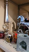 Full view of metal and glass obelisk, the composite horse figurine, black and gold paperweight, glass and metal dishes, and carved stone candle holder all placed together on shelf