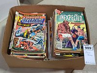 Overhead view of cardboard box containing two stacks of assorted comic books; left stack front shows Marvel Adventure featuring Daredevil, right shows The Unexpected.