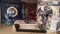 Photo shows a collection of Dungeons and Dragons books, Wizardology book, dragon figurine holding sword, dragon head letter opener, and MEWT cards.
