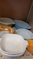 Multiple vintage glass bakeware dishes including Pyrex, Corning Ware, and Anchor Hocking pieces, displayed on a shelf.