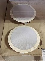 Pair of round Nuvo in-wall speakers showing white metal grills and beige mounting frames, placed on a beige surface.