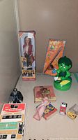 Photo showing a variety of toys including a Barbie doll in box, Green Giant flashlight, handheld football game, Batman figurine, small trucks, and books.