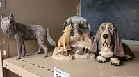 Photo of three figurines on a shelf: a standing ceramic wolf, a resin figurine of two wolves howling at a moon backdrop, and a plaster hound dog lying down.