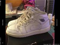 Side profile of right sneaker on black box, showing white leather with creasing and embossed Air Jordan wing logo.