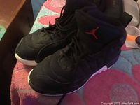 Pair of black Air Jordan Retro 12 kids sneakers showing left side, resting on pink patterned cloth surface with some surrounding items.