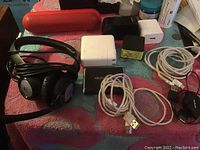 Photo showing a collection of cords, portable chargers, headphones and a red Bluetooth speaker laid on a fabric surface.