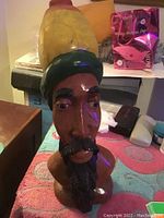 Front view of the hand carved wooden statue depicting a Jamaican Rasta man with detailed facial features, beard, and multicolored hat, placed on a cloth surface with various items in the background.