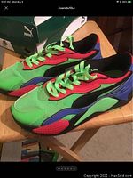 Side angled view of Puma sneakers showing vibrant green, red, black, and blue colors with lime green laces, placed on a wooden surface with Puma shoe box in background.