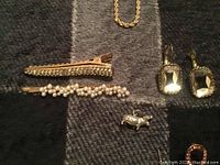 Photo showing various jewelry items on a dark plaid fabric. Two gold-tone hair pins with pearl and rhinestone accents, gold-tone chain necklace, rectangular stone earrings, and a small pig charm visible.