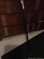 Full length photo of black cane with detailed gun-shaped handle resting against wooden dresser and carpet.