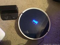 Image showing top view of Strata Home round robot vacuum with digital display glowing blue, placed next to a black charging dock on carpet.