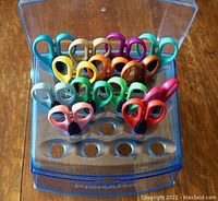 Photo of 13 pairs of Fiskars edging scissors arranged in a transparent plastic holder, showing colorful handles and blade tips.