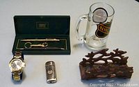 Photo showing two Cross pens (gold-filled and matte black), gold-tone keychain, Bell & Rose wristwatch with metal bracelet, German-made trench lighter, vintage cast-iron letter holder, and Boston Bruins beer mug.