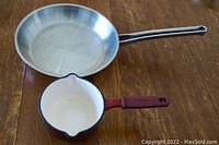 Two cookware pieces on wooden surface: Noser Inox stainless steel frying pan and small Kitchen Aid enameled cast iron sauce pot with maroon handle.