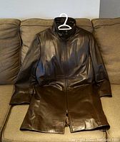 Front view of the brown Danier leather women's coat, laid flat on a sofa, showing the zipper front closure and high collar design.